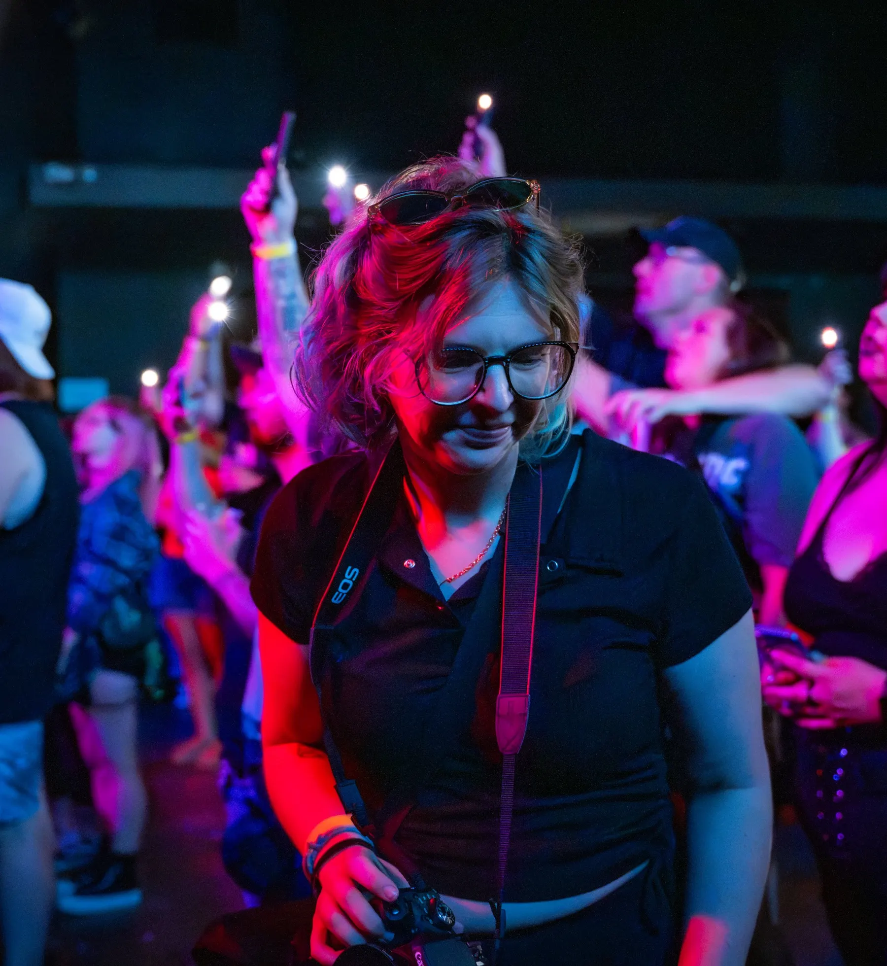 Johnnye in the crowd at a concert, camera in hand, lit pink and blue by the stage lights