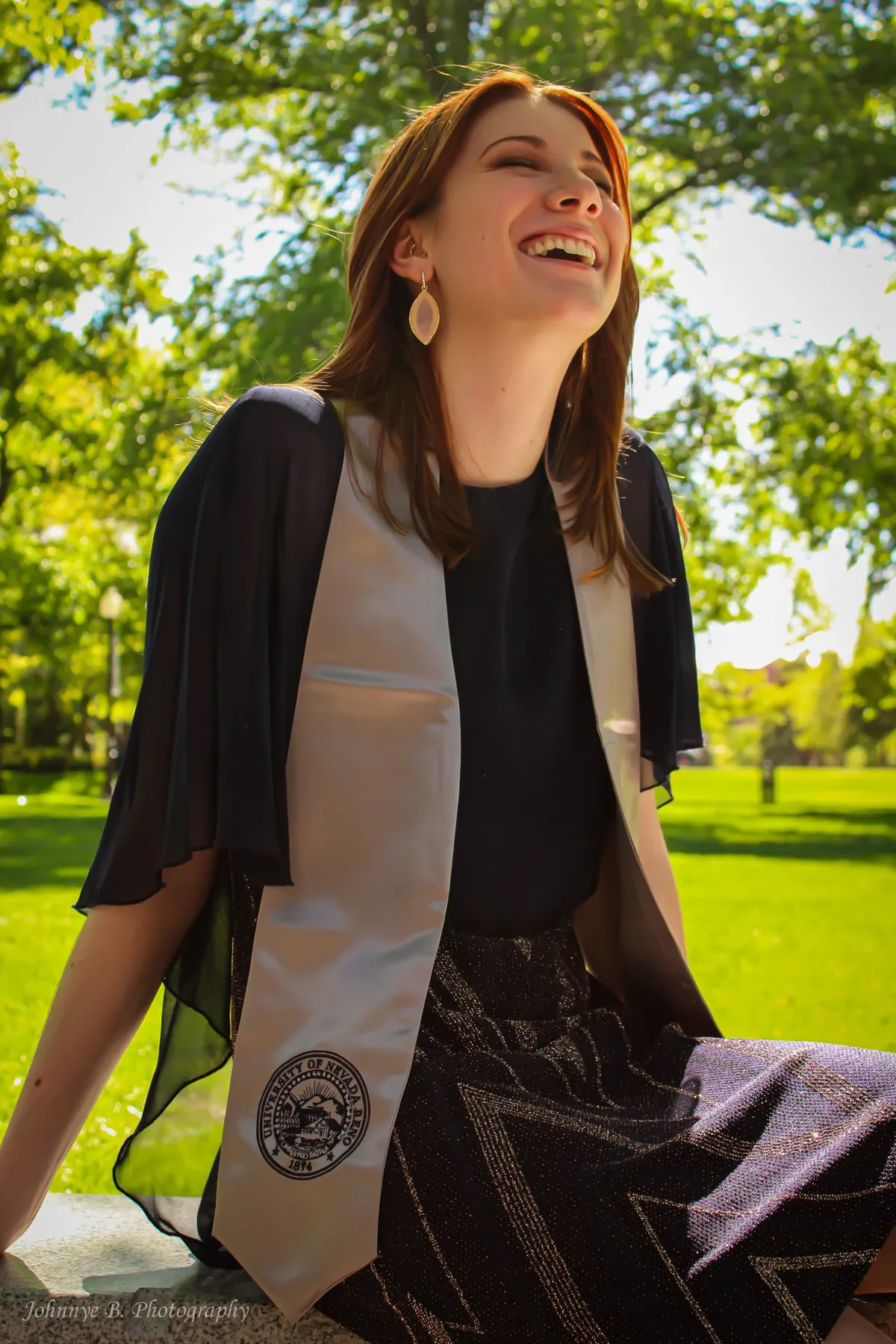 Graduate laughing in cap-and-gown stole on a sunlit campus lawn