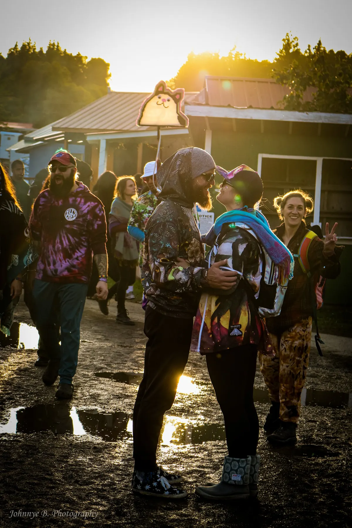 Couple face to face on a muddy festival path at golden hour, a friend throwing a peace sign behind them